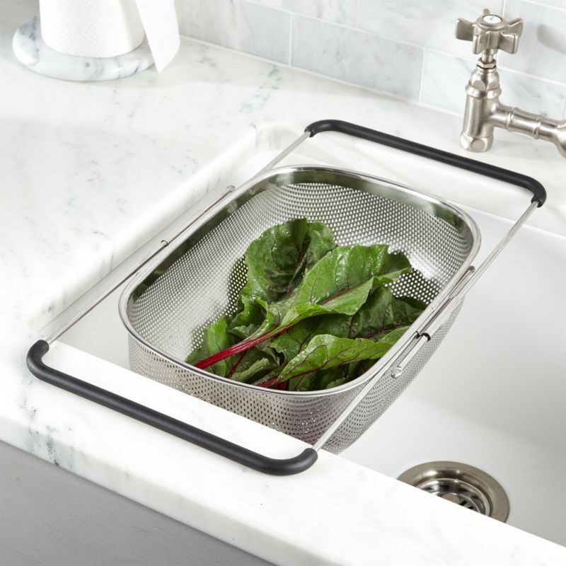 a stainless steel mesh colander resting across a white sink on two black-tipped extending arms, with washed chard in it